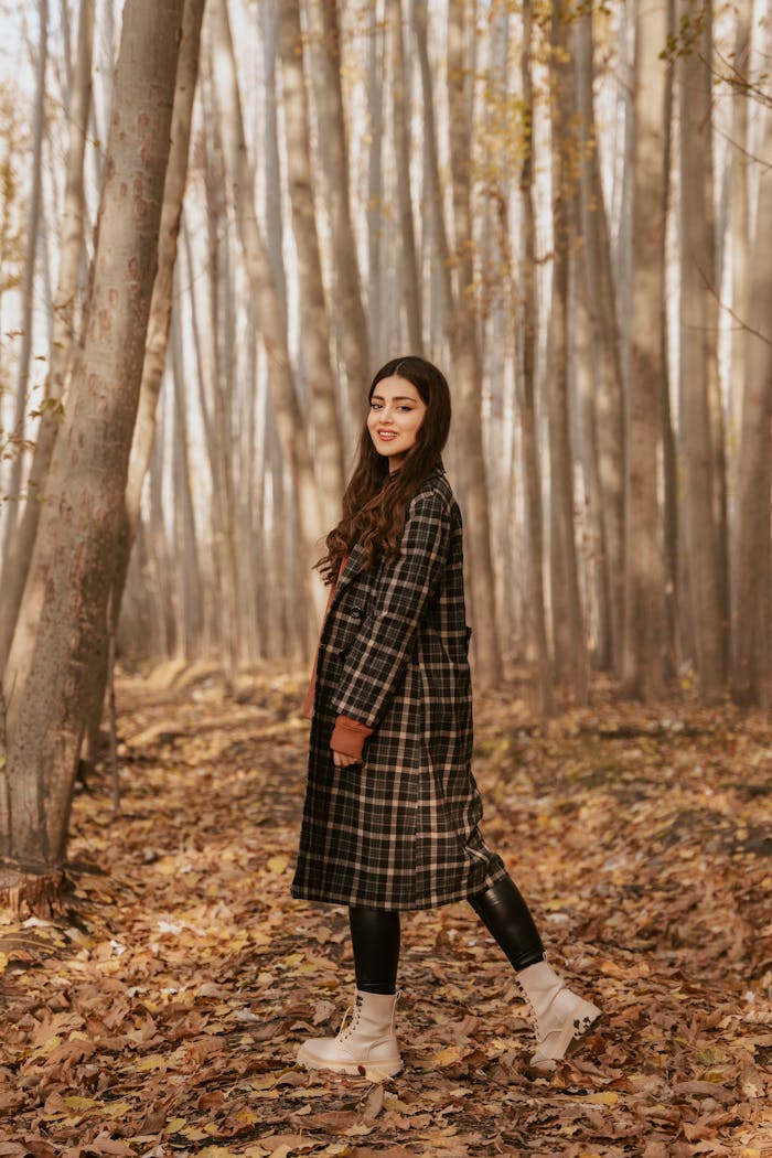 Young woman in plaid coat walking through a forest filled with fallen leaves, smiling and enjoying the autumn setting.