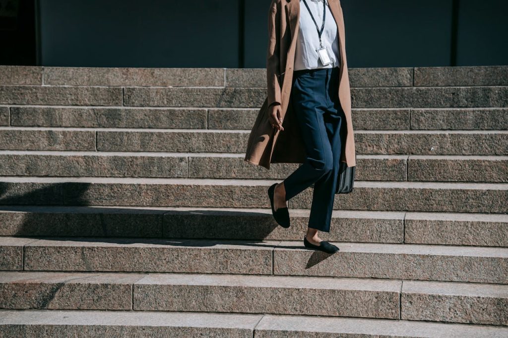 pexels photo 5999966 Crop anonymous female entrepreneur in brown coat with badge and handbag walking down stairs